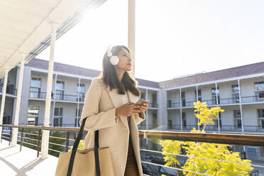 Junge Frau mit Kopfhörern und Smartphone auf dem Balkon - AFVF05364