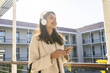 Lächelnde junge Frau mit Kopfhörern und Smartphone auf dem Balkon - AFVF05365