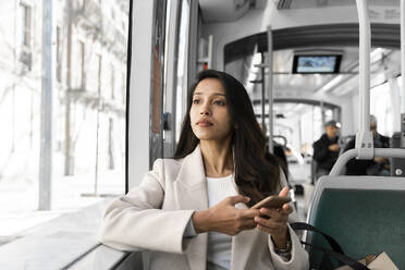 Junge Frau mit Smartphone schaut in einer Straßenbahn aus dem Fenster - AFVF05373