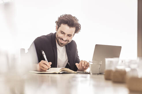 Young man in business jacket working on a laptop and taking notes in a beach house - SDAHF00149