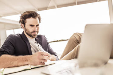 Young man in business jacket working on a laptop and taking notes in a beach house - SDAHF00151