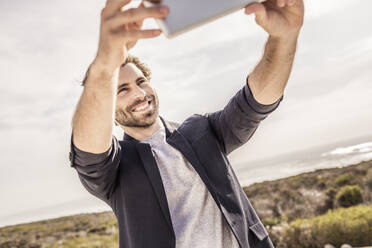 Glücklicher junger Mann in Business-Jacke macht ein Selfie an der Küste - SDAHF00154