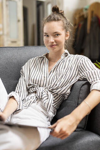 Junge Frau sitzt auf einer Couch, hält ein digitales Tablet und schaut in die Kamera, lizenzfreies Stockfoto