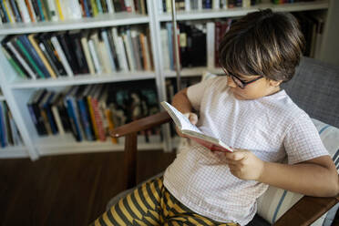 Junge mit Brille sitzt auf einem Sessel und liest ein Buch - VABF02624