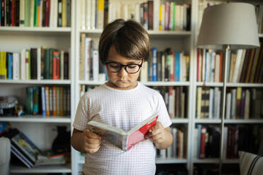Porträt eines ernsten Jungen mit Brille, der vor einem Bücherregal steht und einen Comic liest - VABF02626