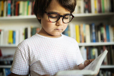 Porträt eines ernsten Jungen mit Brille, der vor einem Bücherregal steht und einen Comic liest - VABF02627