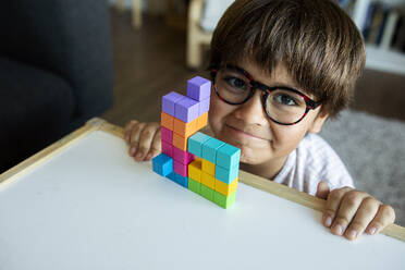 Portrait of smiling little boy with glasses playing with building blocks at home - VABF02643