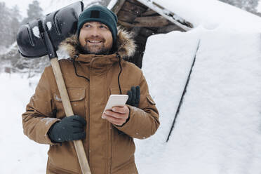 Portrait of smiling man with snow shovel and cell phone - KNTF04405