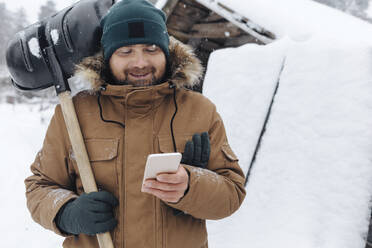 Portrait of smiling man with snow shovel looking at cell phone - KNTF04406