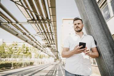 Junger Mann mit Smartphone und Blick zur Seite, in der Stadt - KNSF07593