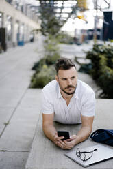 Young man with smartphone and laptop lying on a bench in the city - KNSF07594