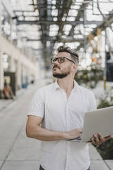 Junger Mann mit Laptop in der Hand und Blick nach oben in die Stadt - KNSF07595