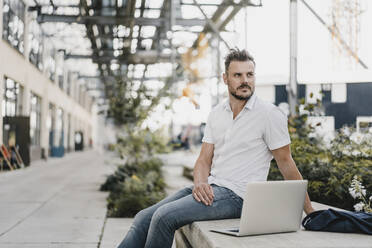 Junger Mann mit Laptop und Blick zur Seite, sitzend auf einer Bank in der Stadt - KNSF07596