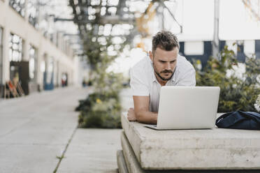 Junger Mann mit Laptop und Blick zur Seite, liegend auf einer Bank in der Stadt - KNSF07597