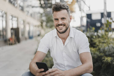 Young smiling man using smartphone and looking at camera - KNSF07599