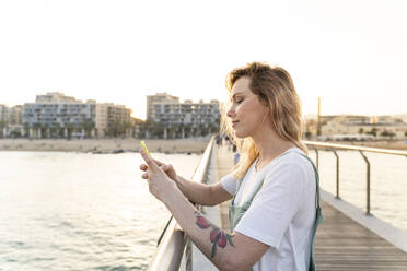 Young woman spending a day at the seaside, using smartphone, standing on bridge - AFVF05415