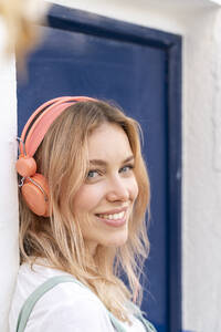 Porträt einer glücklichen jungen Frau, die an einer Fassade lehnt und mit Kopfhörern Musik hört - AFVF05423