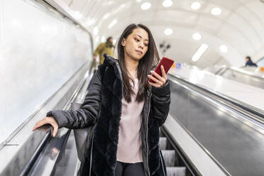 Porträt einer Frau, die auf einer Rolltreppe steht und auf ihr Mobiltelefon schaut, London, UK - WPEF02618