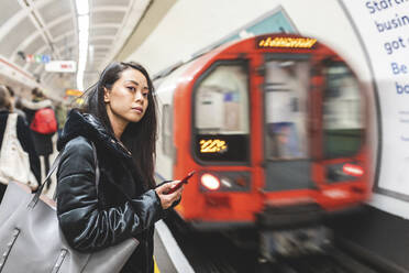 Portrait of woman with smartphone waiting at underground station platform, London, UK - WPEF02619
