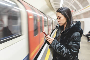 Porträt einer Frau, die am Bahnsteig einer U-Bahn-Station wartet und auf ihr Smartphone schaut, London, Vereinigtes Königreich - WPEF02620