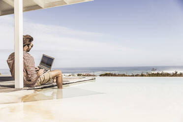 Mann arbeitet am Laptop neben dem Pool in einem Strandhaus - SDAHF00159