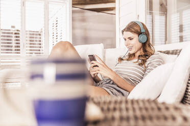 Junge Frau entspannt sich mit Smartphone und Kopfhörern auf einer Veranda - SDAHF00179