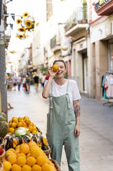 Porträt einer lachenden jungen Frau in einer Einkaufsstraße, die ihr Auge mit einer Orange bedeckt - AFVF05450