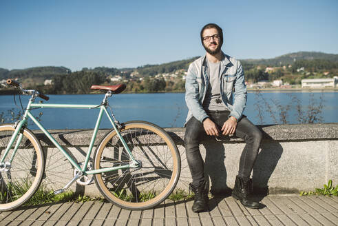 Junger Mann auf einer Mauer am Meer sitzend, mit seinem Fixie-Fahrrad - RAEF02335