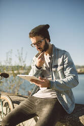 Junger Mann sitzt am Meer und benutzt ein digitales Tablet - RAEF02336