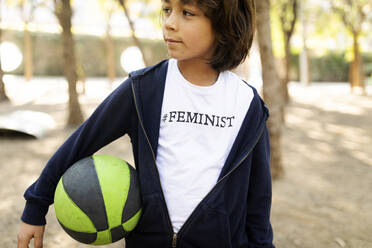 Little boy standing in the street with print on t-shirt, saying Feminist, holding ball - VABF02646