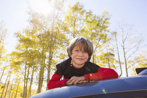 Ein lächelnder sechsjähriger Junge liegt auf der Motorhaube eines blauen Geländewagens im Wald., lizenzfreies Stockfoto