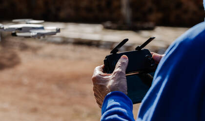 Rear view of the elderly man piloting the drone looking at the remote control display while standing - CAVF74690
