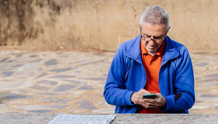 Senior man looking and touching screen of smartphone while sitting on park - CAVF74694