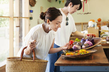 Zwei lächelnde Japanerinnen sehen sich in einem Hofladen frisches Gemüse an. - MINF13694