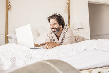 Mann im Bademantel, auf dem Bett im Schlafzimmer liegend, mit Laptop - SDAHF00289