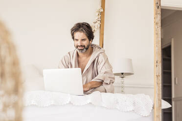 Man wearing bathrobe and sitting on bed in bedroom - SDAHF00290