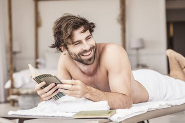Young man with beard reading a book, lying on lounge - SDAHF00292