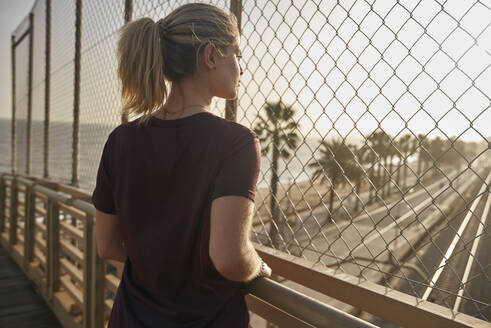 Sportliche junge Frau steht auf einer Brücke und schaut durch einen Zaun - PACF00195