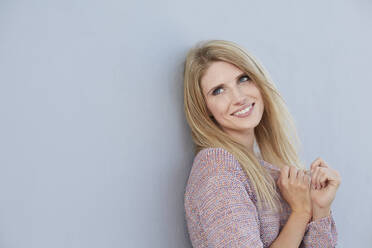 Porträt einer lächelnden blonden jungen Frau, die einen Strickpulli trägt und sich an eine Wand lehnt - PNEF02346
