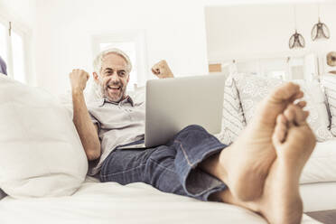 Mature man with laptop cheering on couch at home - SDAHF00330