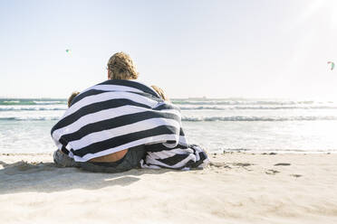 Rückansicht eines Vaters mit zwei in ein Handtuch gewickelten Söhnen am Strand - SDAHF00347