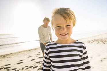 Porträt eines lächelnden Jungen am Strand mit Vater im Hintergrund - SDAHF00350