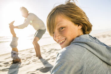 Porträt eines lächelnden Jungen mit einem Vater, der seinem Sohn bei einem Kopfstand am Strand im Hintergrund hilft - SDAHF00354