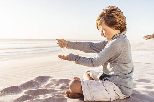 Kleiner Junge sitzt am windigen Strand und beobachtet, wie der Sand von seinen Händen rieselt - SDAHF00408