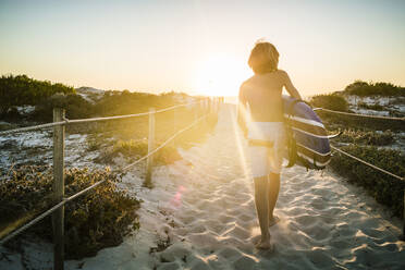 Rückansicht eines Jungen mit Surfbrett, der bei Sonnenuntergang zum Strand geht - SDAHF00425
