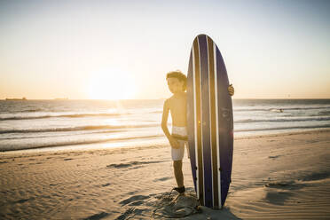 Junge mit Surfbrett steht am Strand bei Sonnenuntergang - SDAHF00428