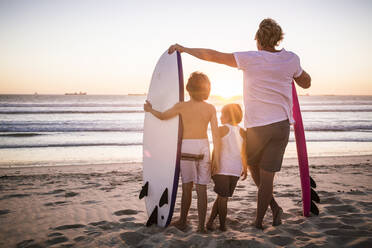 Rückansicht von Vater und Söhnen mit Surfbrettern, die bei Sonnenuntergang am Strand stehen - SDAHF00432