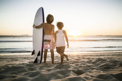 Rear view of two boys with surfboard standing on the beach at sunset - SDAHF00433