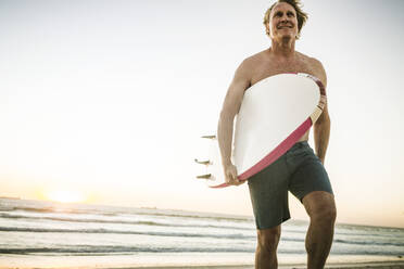 Lächelnder Mann mit Surfbrett am Strand bei Sonnenuntergang - SDAHF00434