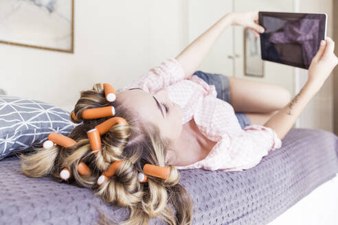 Junge Frau mit Lockenwicklern im Haar liegt auf dem Bett und macht ein Selfie mit einem digitalen Tablet - SDAHF00439